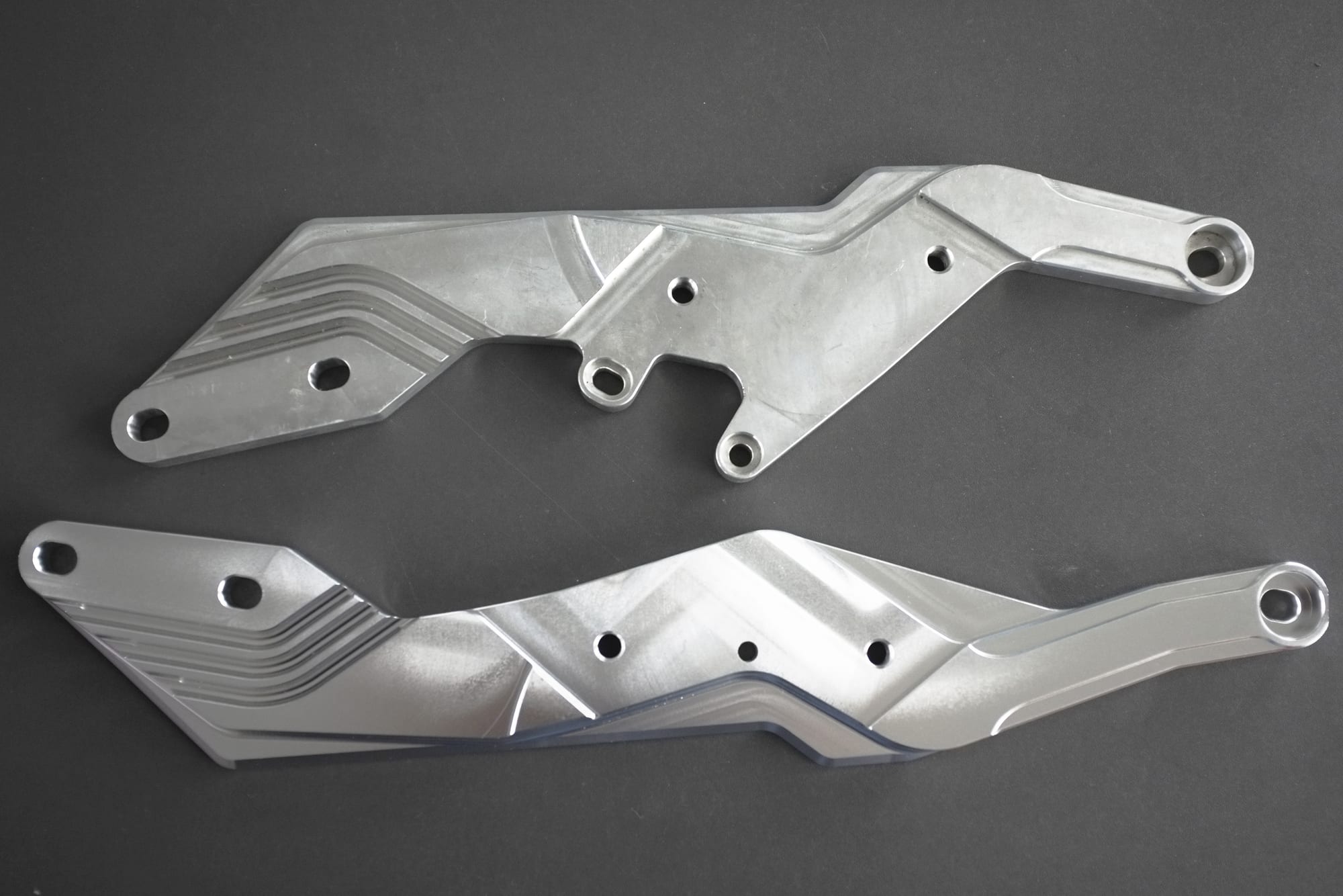 The image shows a pair of aluminum rear lower control arms for a Yamaha YZF-R1 on a black surface. The metal parts are silver in color and have a sleek, modern design.
